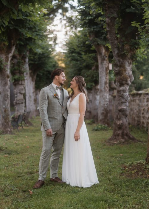 Alina_Marc-361 Eine Braut und ein Bräutigam stehen zusammen im Freien in einem üppigen, grünen Garten, umgeben von hohen Bäumen. Der Hochzeitsfotograf fängt ihr Lächeln ein, während der Bräutigam einen hellgrauen Anzug trägt und die Braut in ihrem ärmellosen weißen Kleid strahlt.