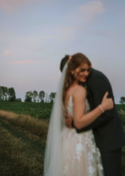 Hochzeit (1107) Eine Braut in einem weißen Kleid und Schleier umarmt ihren Bräutigam im Freien bei Sonnenuntergang, grüne Felder und Bäume im Hintergrund. Beide Figuren sind leicht unscharf, was diesem Hochzeitsfotograf-Bild eine verträumte, intime Atmosphäre verleiht.