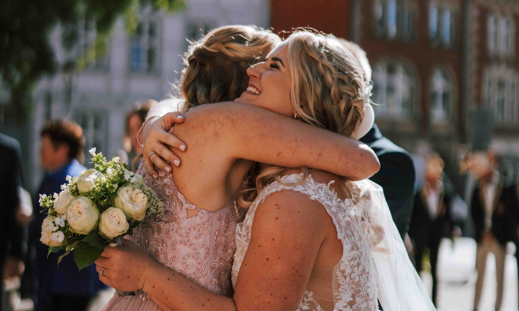 Hochzeit (443) Die Braut in ihrem schönen weißen Kleid mit Blumen und Brautstrauß umarmt eine Freundin in einem schönen rosa Kleid bei der Hochzeit in Duderstadt in der Innenstadt mit Ringen an den Händen. Foto von der Fotografin.