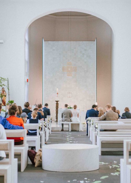 Hochzeit (95) Eine Gruppe von Menschen sitzt auf weißen Bänken in einer modernen, hellen Kirche und blickt auf den Altar. Der Raum ist minimalistisch mit weißen Wänden, hohen Fenstern und einer einzigen hohen Kerze - perfekt für jeden Fotografen, der eine Hochzeit festhält.
