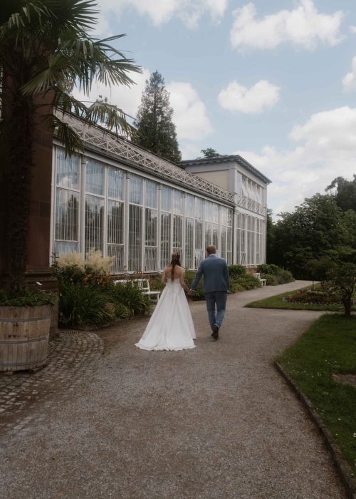Hochzeit-Lena-Marcel-1 Braut und Bräutigam spazieren Hand in Hand über einen Gartenweg neben einem großen Glaswintergarten, umgeben von Grün und unter einem teilweise bewölkten Himmel - perfekte Momente für jeden Hochzeitsfotografen, um sie an Ihrem besonderen Tag festzuhalten.