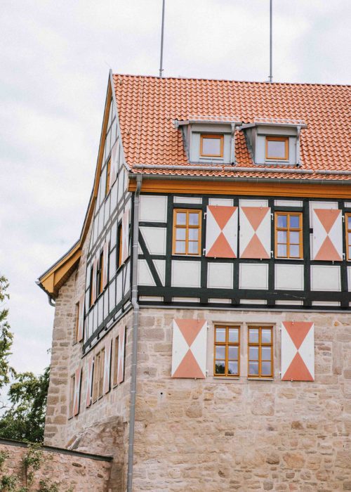 Hochzeit9 Ein Fachwerkhaus mit rotem Ziegeldach, beigen Steinwänden und dekorativen roten und weißen Fensterläden - eine ideale Kulisse für eine Hochzeit oder Trauung. Der wolkenverhangene Himmel und das Grün auf der linken Seite verleihen dem Bild einen ganz besonderen Charme, perfekt für die Linse eines Fotografen.